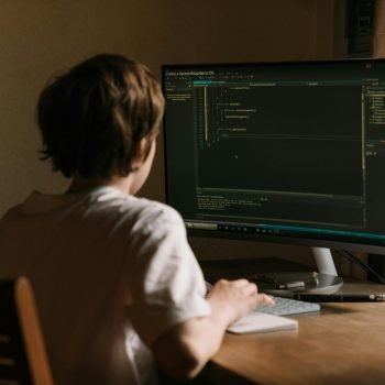 A teenager focused on coding software on a desktop monitor in a home office setting.